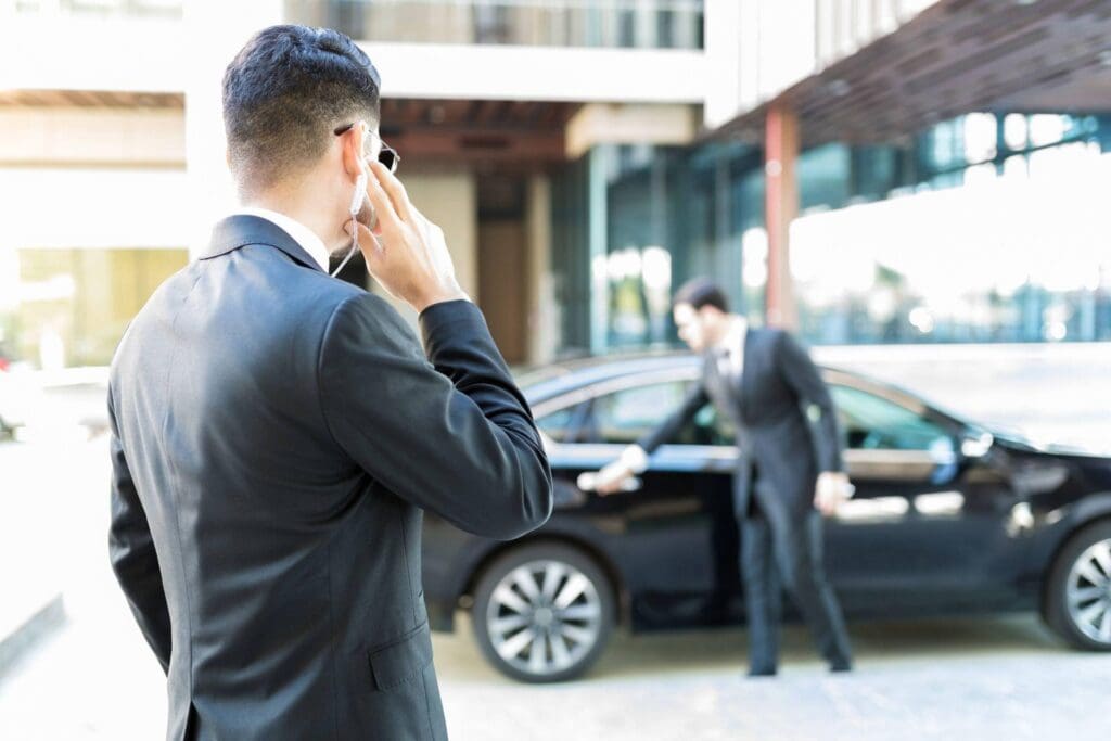 Man in a suit using an earpiece while another man opens a black car door